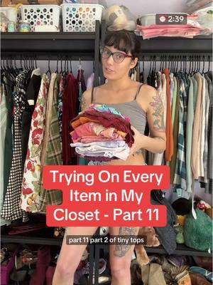 Trying on Every Item in my Closet - Part 11 - Part 2 of Tiny (Summet) Tops #ClosetTour #TryOnHaul #StyleInspo #ManicChangeling #ClosetCleanOut #OpenConceptCloset #ThriftedFashion #Thrifting #CropTop #CropTopStyle  Video length: 2 minutes 39 seconds. Video description: Liddi, a slim person with tan skin, colorful tattoos, and black hair tied back except for bangs is speaking to the camera while holding a stack of folded shirts. She is wearing glasses. She tries on each of the tops with verbal description and spins around to show the full item. 