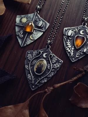 These handcrafted shields, weathered and battle-worn, are intended to guard the heart of the wearer and guide them toward triumph. All handmade from sterling silver, they each have their own unique qualities.  🛡️ The first shield, divided into four quadrants, features a natural faceted orange garnet in the center, seated within solid 18k gold. A brass sun, sterling pumpkin, brass crescent moon, and sterling triple spiral adorn the shield. These symbols call to the cycles of light and dark and the never ending process of becoming, rendering this shield the ultimate armor for shadow work and self-actualization.  🛡️ Upon the second shield, oak leaves and thorny bramble branches unfurl in a secure embrace around a natural dendritic agate, topped with a sterling crescent moon. These plants grant the wearer sharp protection, ancestral strength, and rooted integrity in all matters. This shield is a reminder to stay true to oneself. 🛡️ The third shield bursts with the abundance of pumpkins and wheat, which grow around a natural faceted orange garnet. A crescent moon floats serenely above the stone. This shield represents the inspiration and magic that follow when one stands firm in their convictions.  All three of these pendants were crafted in homage to the fortitude of the human spirit. They each hang from 18” sterling chains (length can be altered upon request), and will be part of The Final Harvest Vol. IV, which debuts on 2/13 at 8pm et! 🖤 #soliloquyjewelry #crystaljewelry #medieval #fantasy #witchy #necklaces #jewelry #fantasyjewerly 